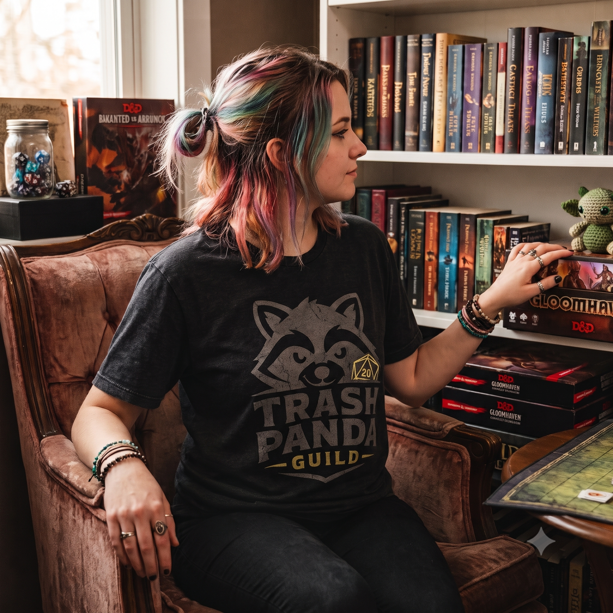 A woman with colorful hair sits in a brown armchair in a room with bookshelves, wearing a black graphic t-shirt that reads 'TRASH PANDA GUILD'.